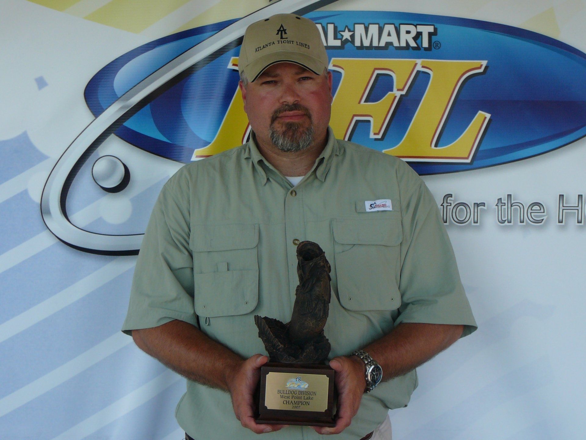 Man in a tan shirt and cap holding a fishing trophy in front of a FLW banner.