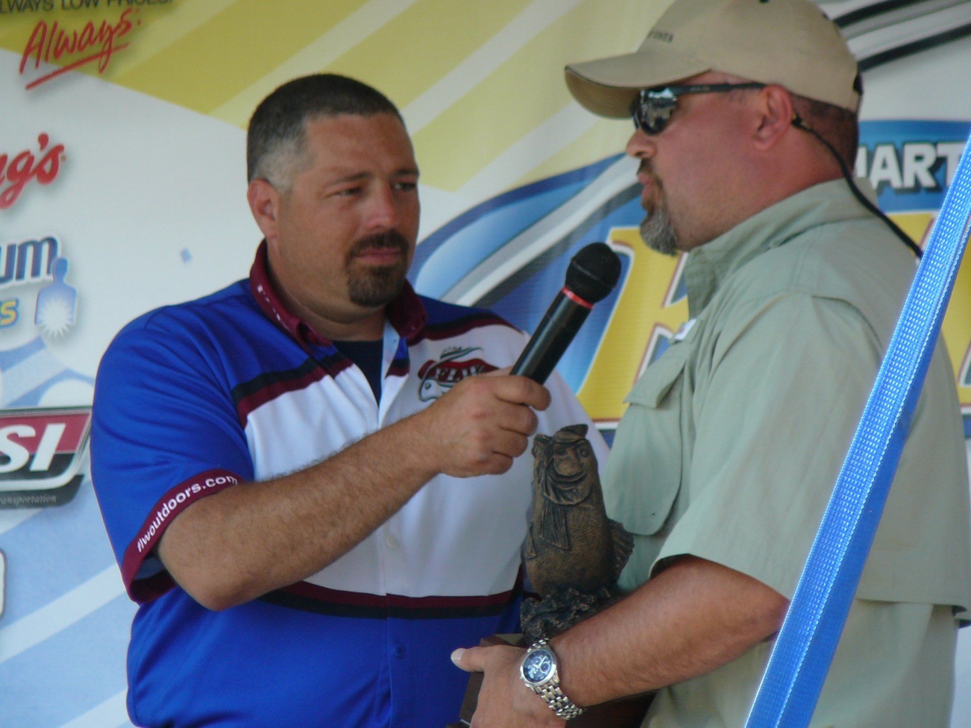 Man in blue shirt interviews man holding trophy. Outdoors, stage setting.