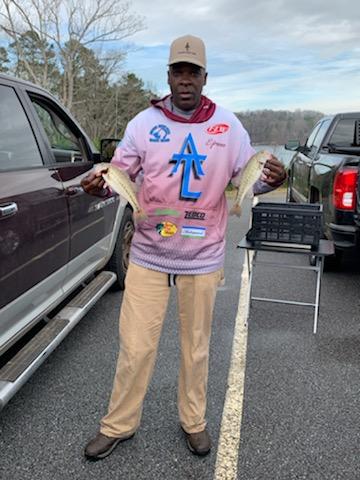 Man holds two fish, standing next to trucks. He wears a shirt with logos, tan pants, and a cap. Outdoors on a cloudy day.