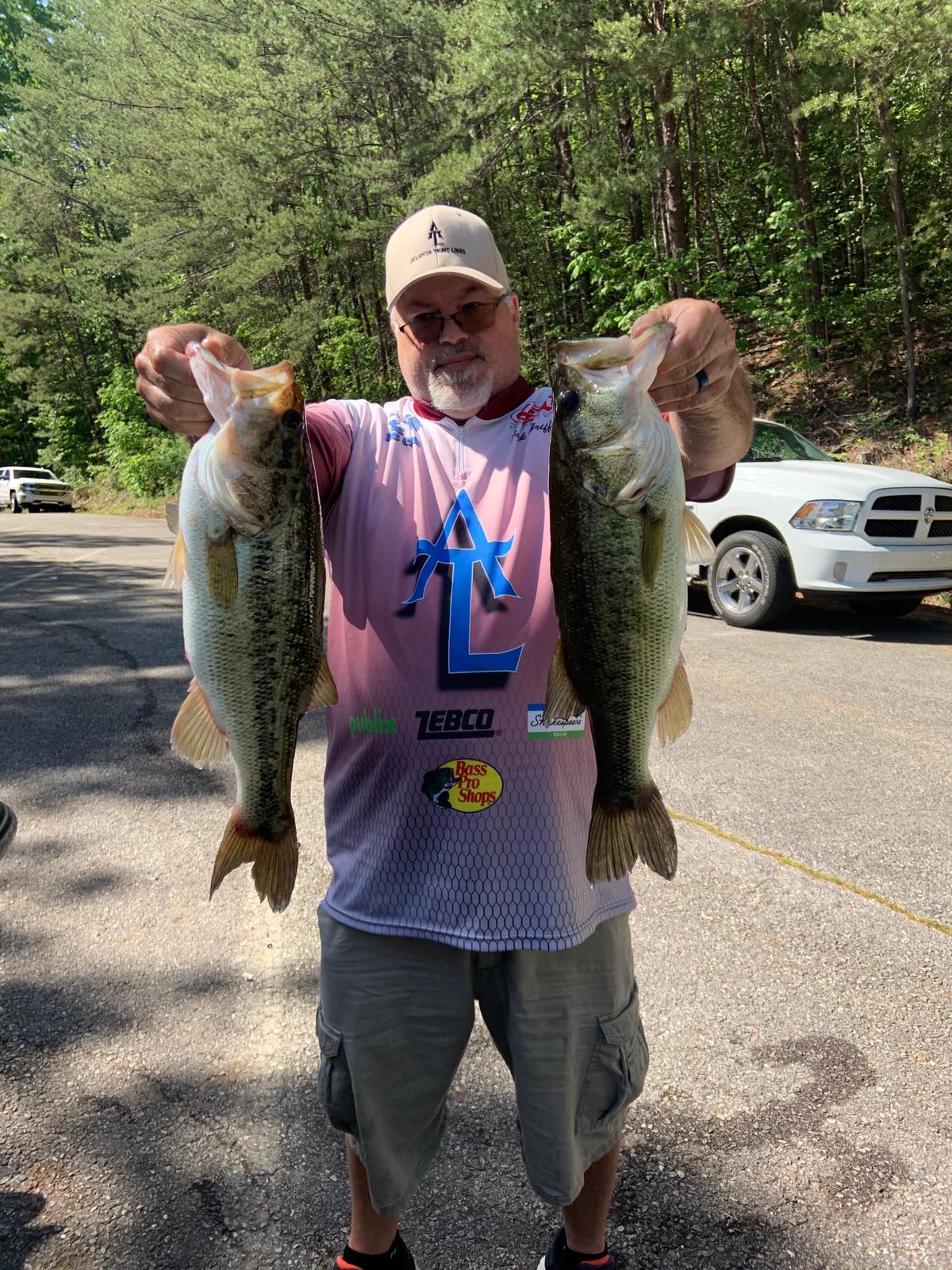 Man holding two large bass, standing on a paved road. White truck in the background.