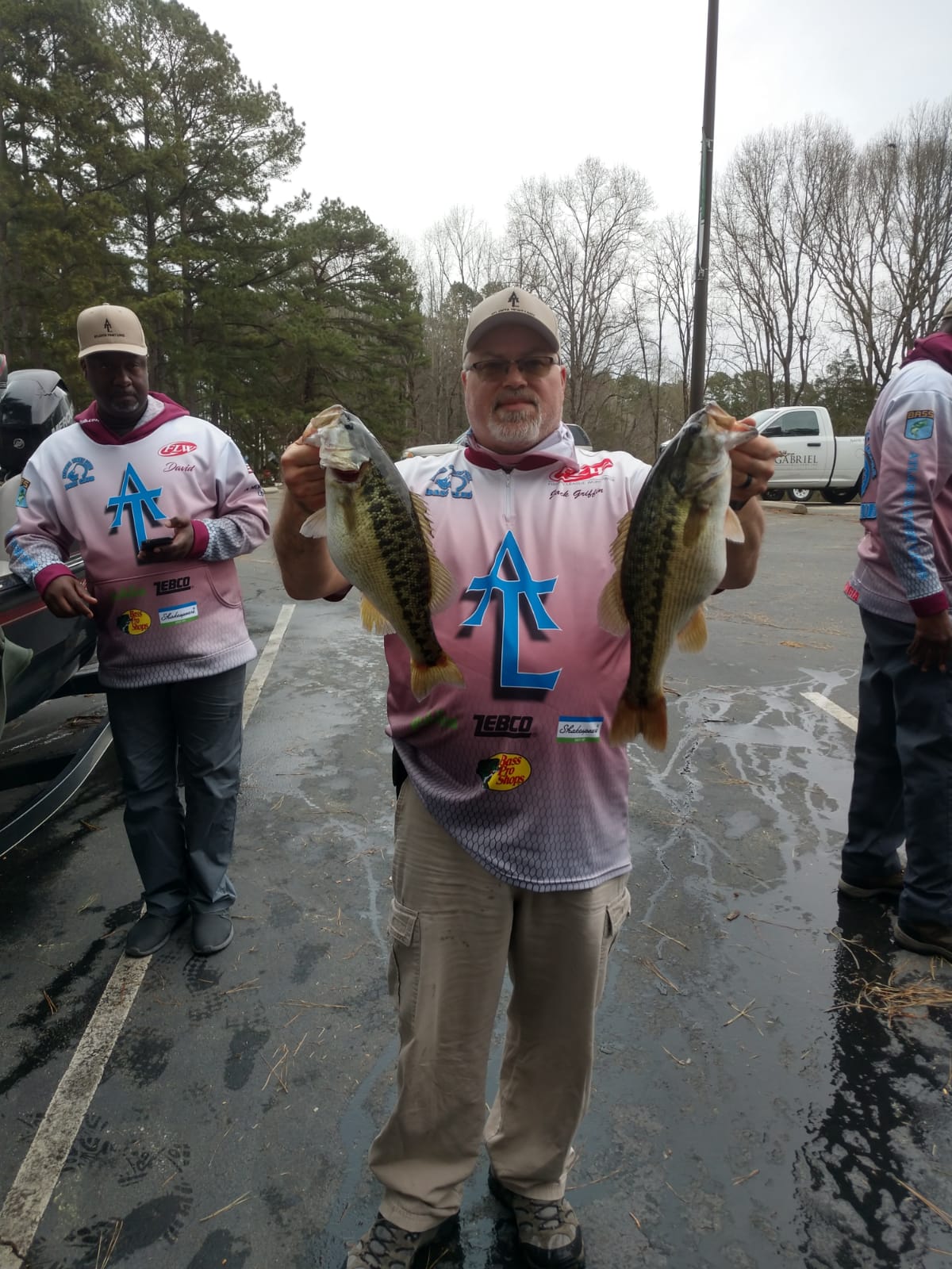 Man holding two large fish, outdoors, overcast day, others in background, wearing fishing apparel.