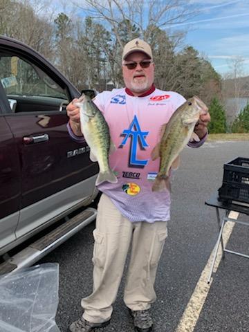 Man holding two largemouth bass near a truck. He wears fishing gear, smiles, and is outdoors.