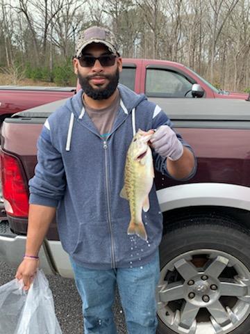Man in blue hoodie holding a fish in front of a red pickup truck; wearing hat, sunglasses, and gloves.
