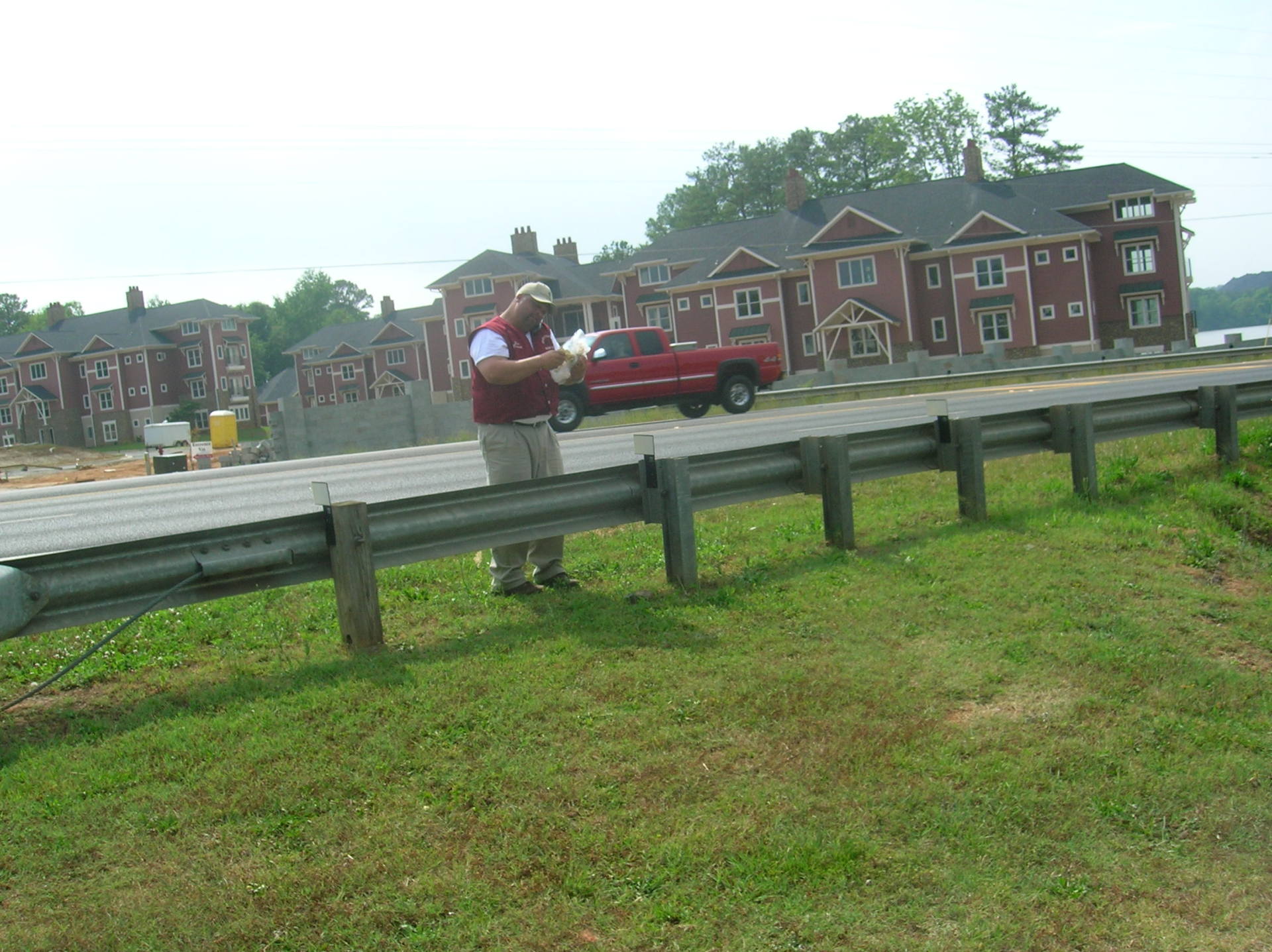 Man standing near guardrail, looking at papers. Red buildings in background. Green grass and sky.