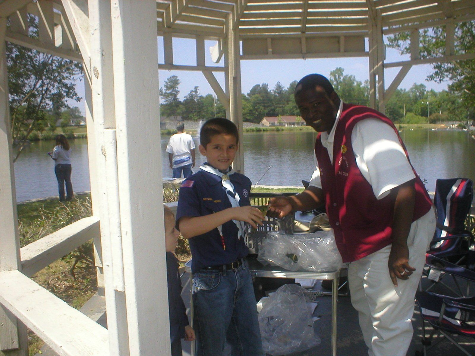 Boy Scout and man smiling at a table by a lake. Man points at something, boy holds item. Outdoors, sunny day.
