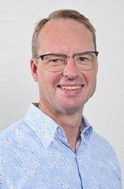 Man with glasses smiles, wearing a light blue patterned button-up shirt, against a white backdrop.