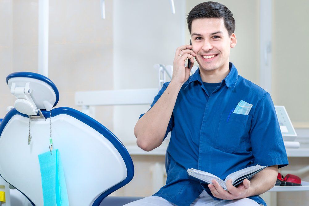 Smiling dentist in blue scrubs on the phone beside a dental chair, holding an open book in a clinic.