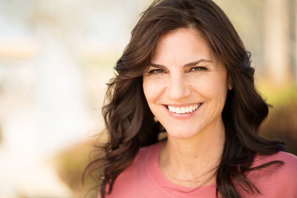 Smiling woman with dark hair wearing a pink top outdoors in soft sunlight