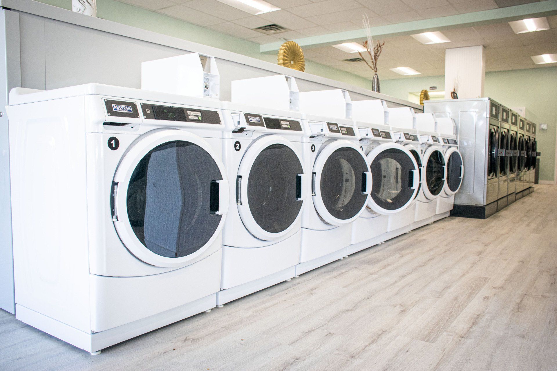 Row of white washing machines in a brightly lit laundromat with light wood-look flooring.