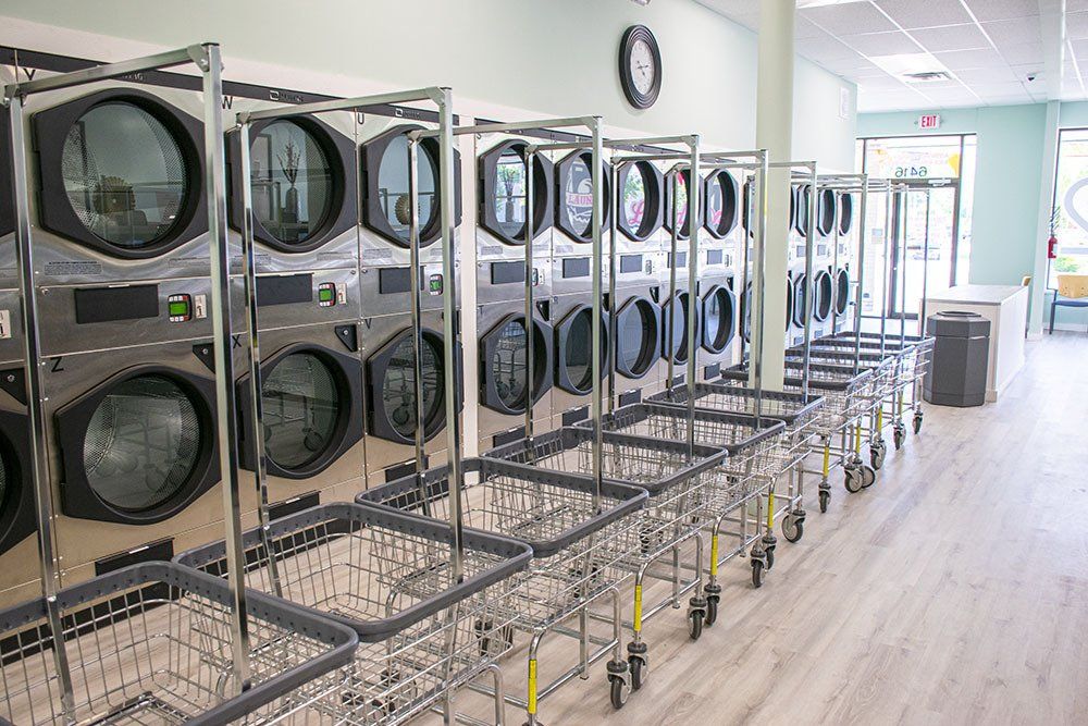 Row of large silver laundry dryers and rolling laundry carts in a brightly lit laundromat.
