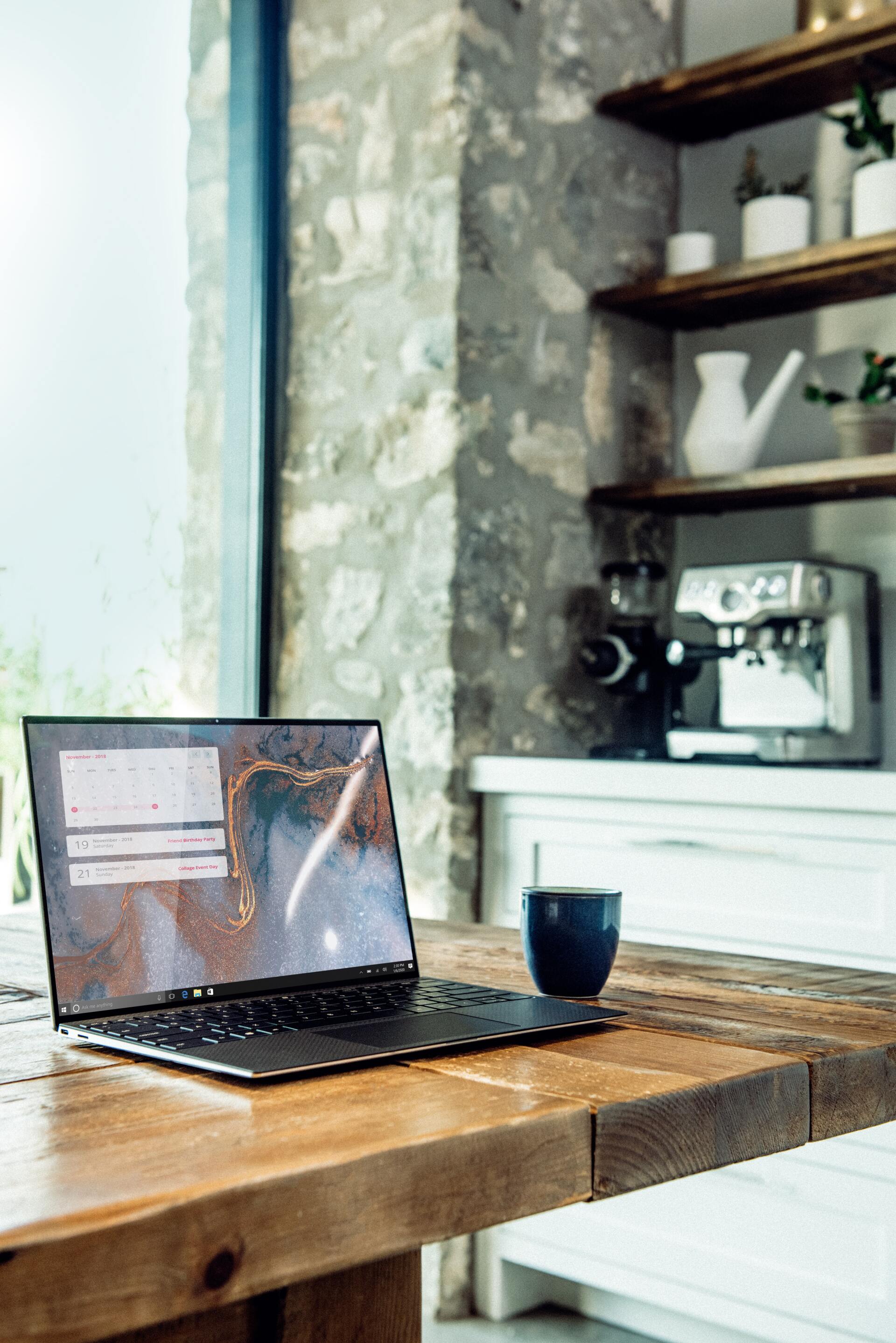 Laptop op een houten tafel met een kop koffie. Keuken met stenen muur en espressomachine op de achtergrond.