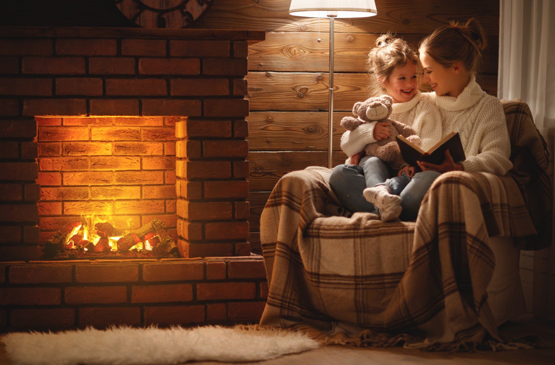 Person and child sitting by a glowing fireplace in a cozy living room, reading together on a plaid chair.