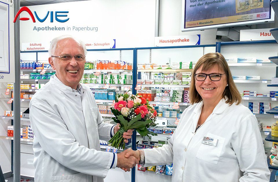Ein Mann und eine Frau geben sich in einer Apotheke die Hand, er gibt ihr einen Blumenstrauß.