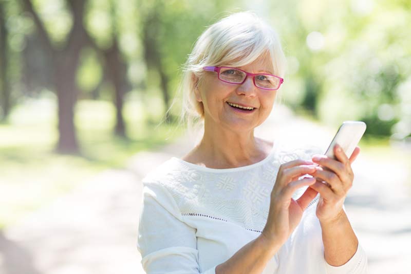 Eine ältere Frau mit Brille benutzt in einem Park ein Mobiltelefon.