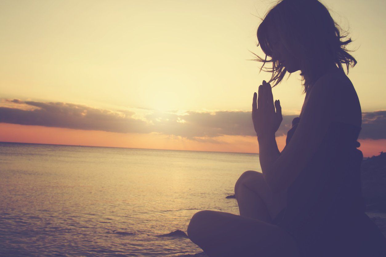 A woman is sitting on the beach with her hands folded in prayer.