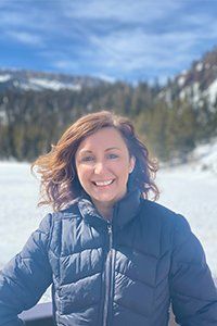 Smiling person in a navy puffer jacket standing in a snowy mountain landscape under a blue sky