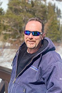 Smiling man in blue jacket and sunglasses outdoors with snowy trees in the background