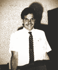 Smiling person in a white shirt and tie standing indoors against a plain wall