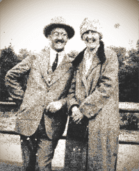 Two people in vintage coats and hats smiling outdoors by a wooden fence