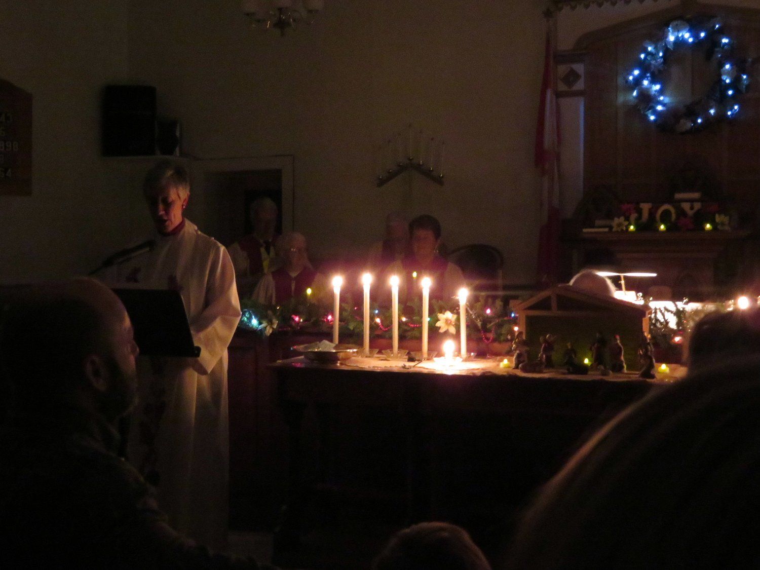 Dimly lit candlelit indoor gathering with people standing near a decorated table and glowing wreath on the wall