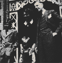 Black-and-white photo of two men on a stage, one standing and one seated beside an ornate backdrop.