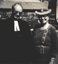 Two people posing outdoors, one in a dark suit and the other in a hat and patterned dress.
