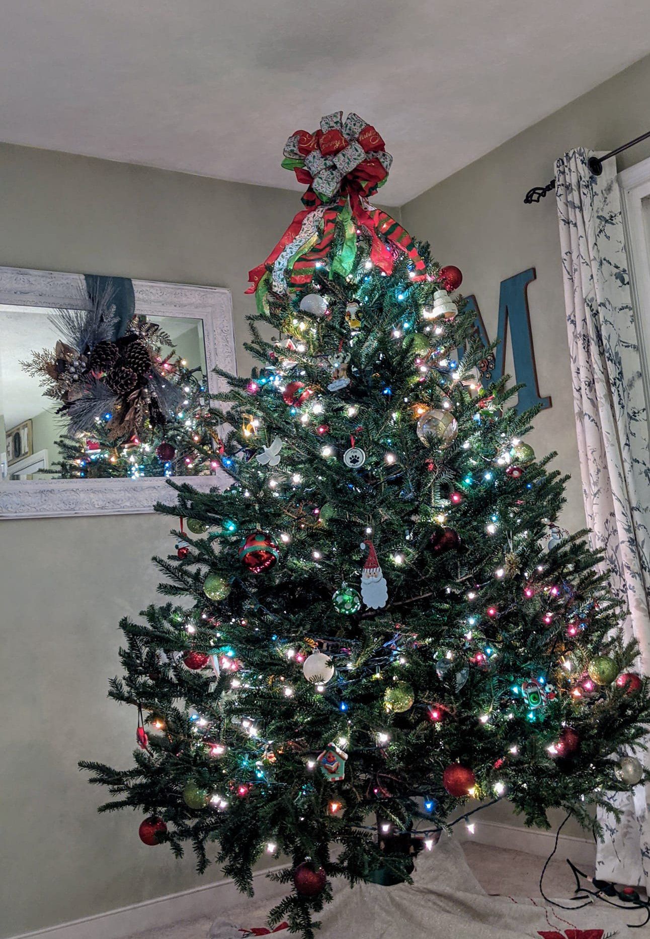 A christmas tree is decorated in a living room in front of a mirror.