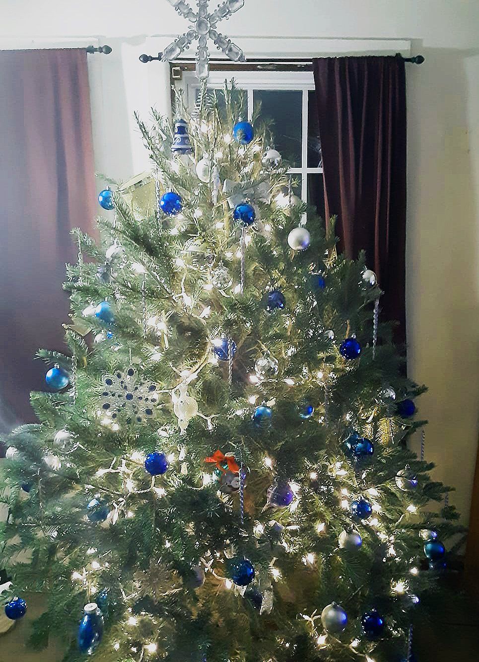 A christmas tree is decorated with blue and silver ornaments and lights in a living room.