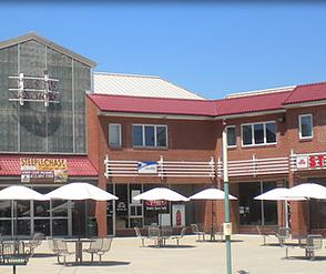 Outdoor mall with brick buildings, red roofs, umbrellas, and tables. Includes a restaurant, post office, and other shops.