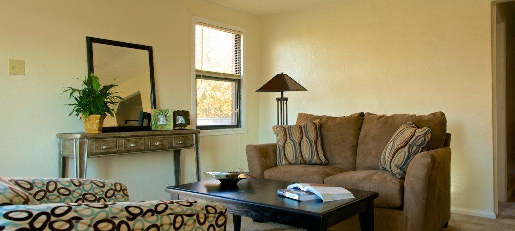 A living room with a brown sofa, coffee table, and decorative console table. Beige walls and natural light.