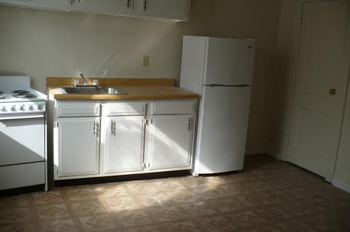 Empty, small white kitchen: sink, cabinets, refrigerator, and stove. Sunlight on floor.