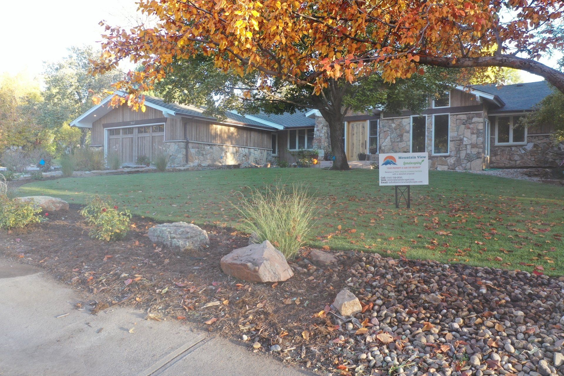 House with stone exterior, lawn, and fall foliage. A sign is in the yard.