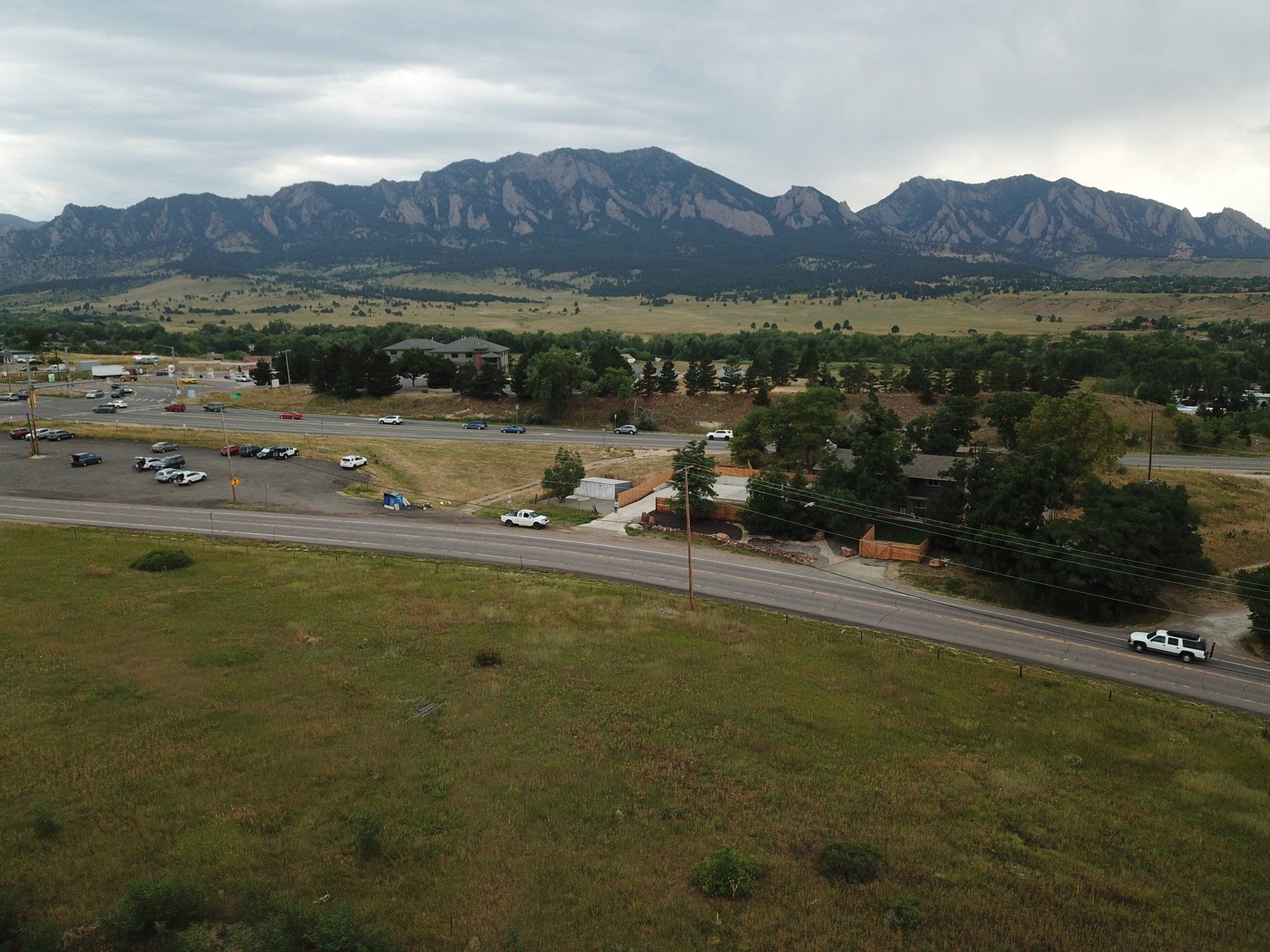 Grassy field leads to a road with cars, buildings, and a mountain range under cloudy skies.