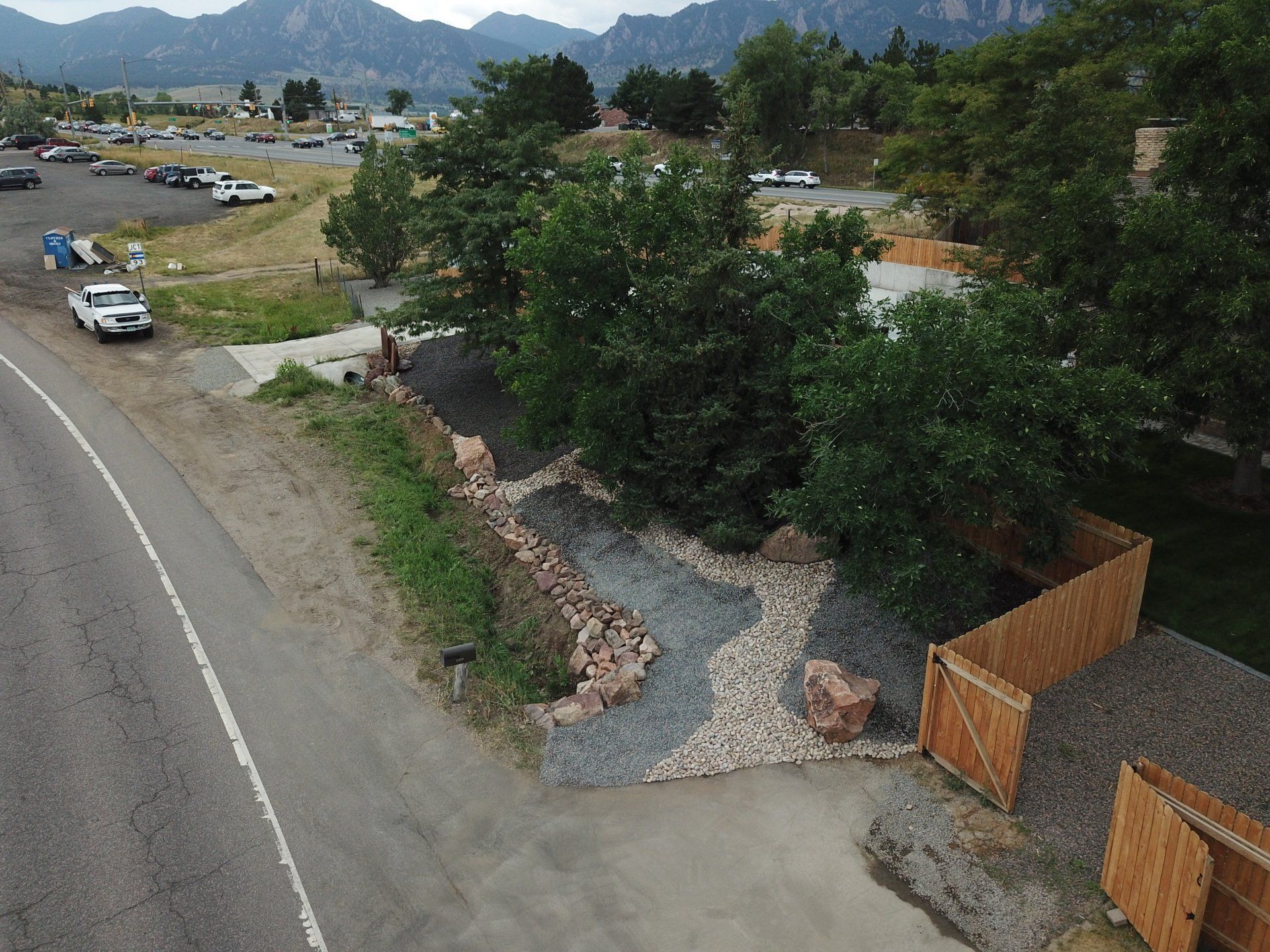 Roadside view: a small park entrance, gray asphalt, gravel paths, trees, parking lot, mountains in the distance.