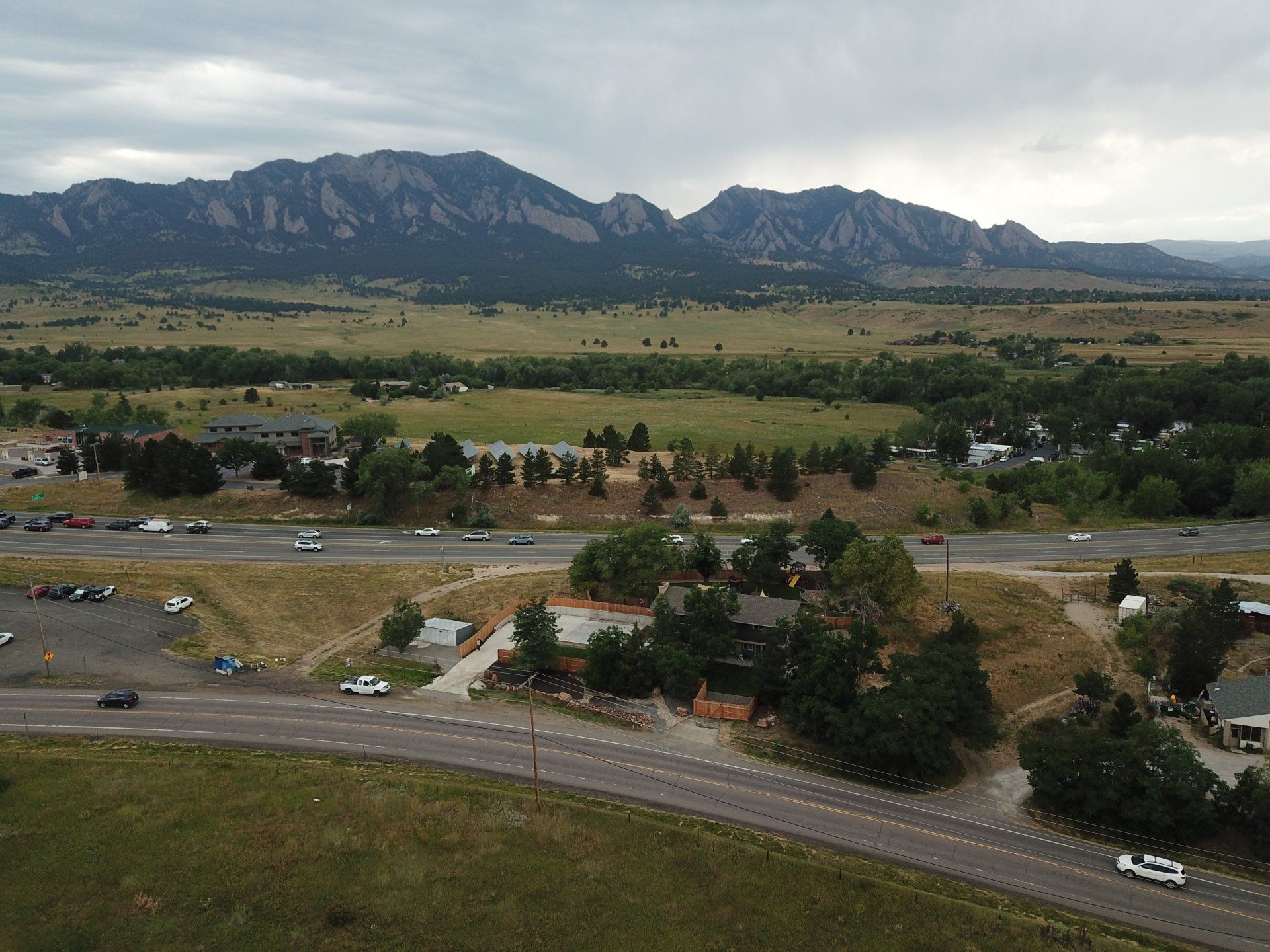 Mountains overlook a highway and green fields; a building with a parking lot in the foreground. Overcast sky.