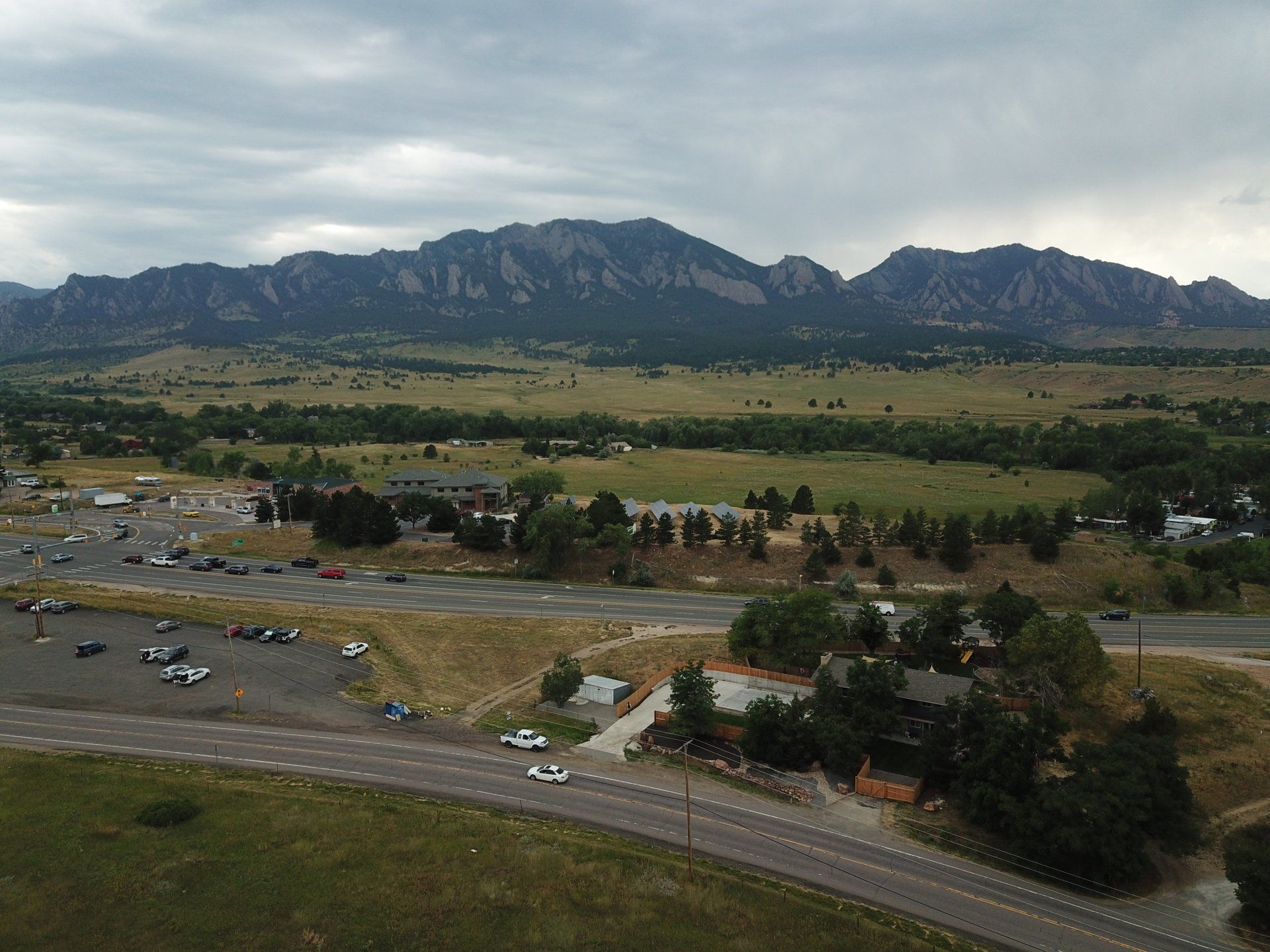 Mountain range backdrop over a grassy plain, a road, parking lots, and scattered buildings. Overcast sky.