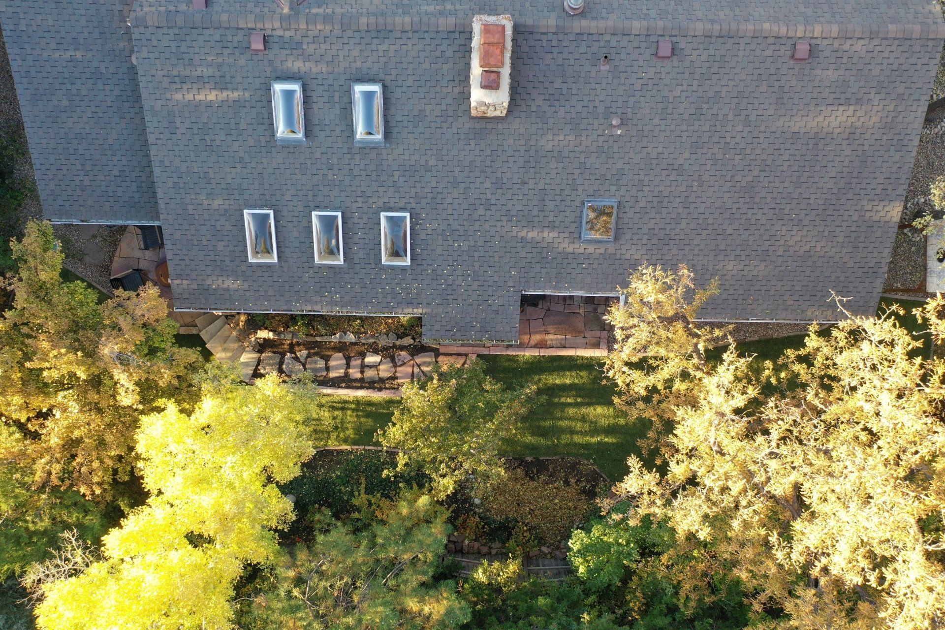 Overhead view of a house with a gray roof, skylights, chimney, and surrounding trees in fall foliage.