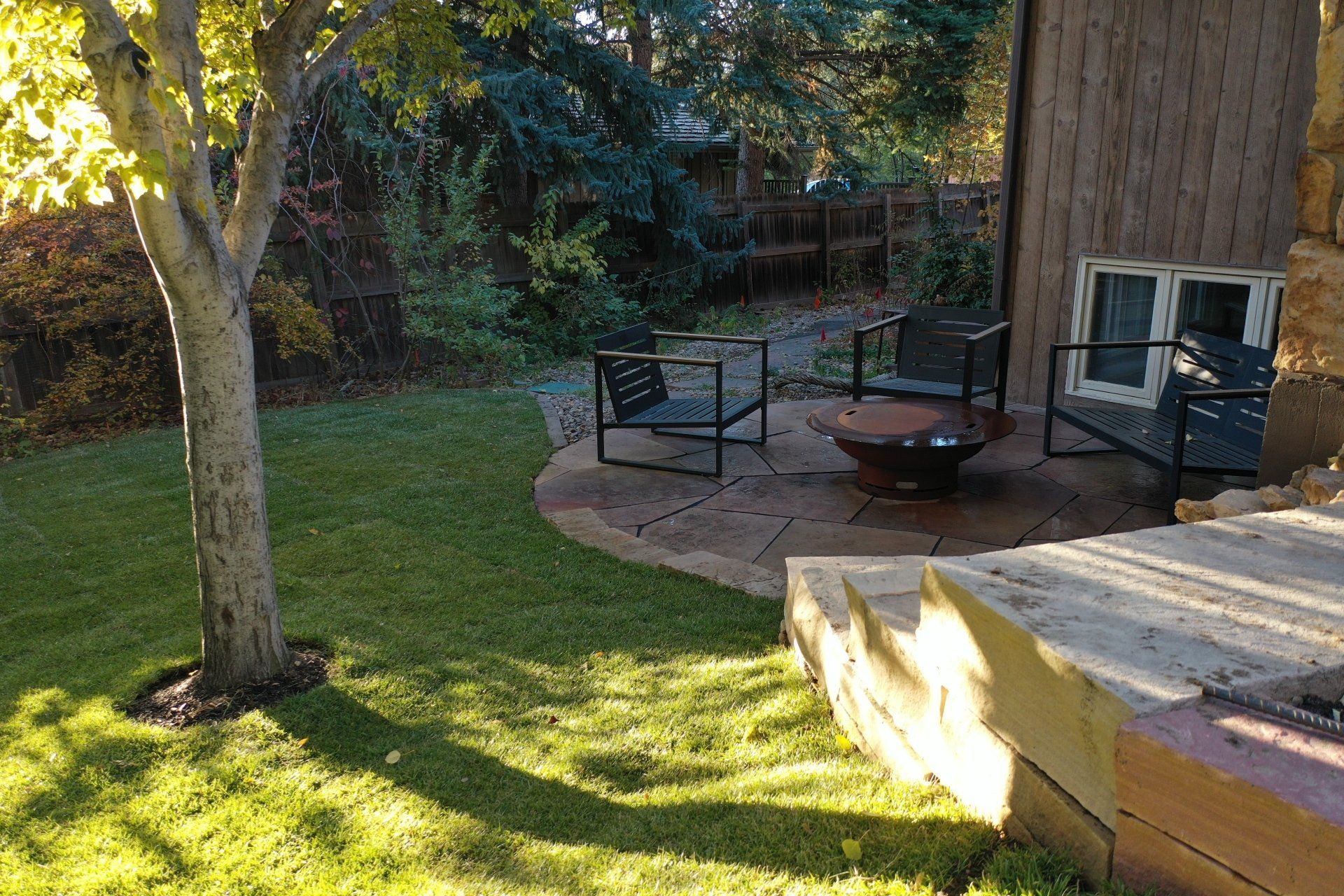Backyard patio with fire pit, chairs, and stone wall, surrounded by green grass and trees.