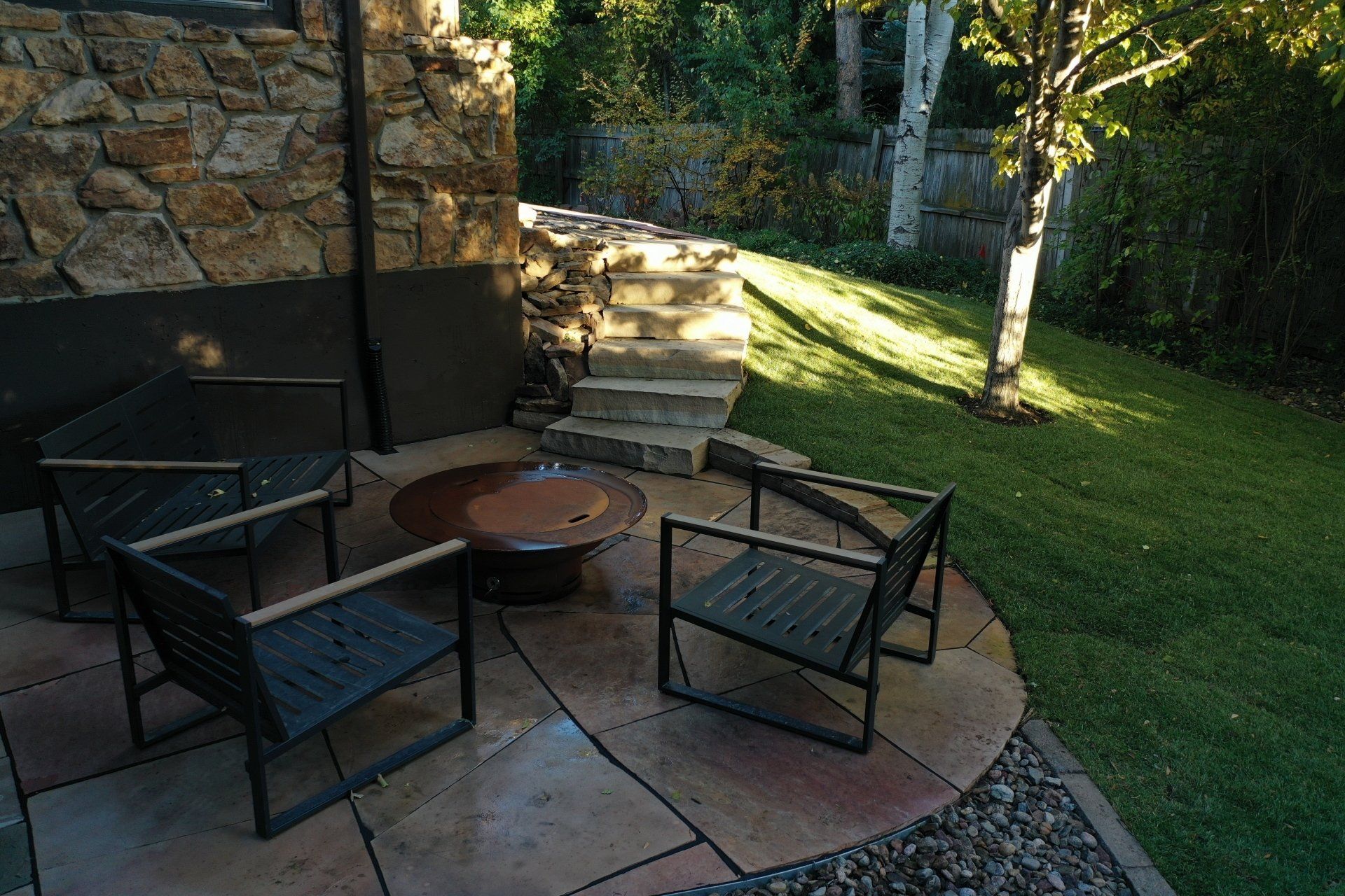 Patio with fire pit, seating, stone wall, steps, tree, and grass in outdoor setting.