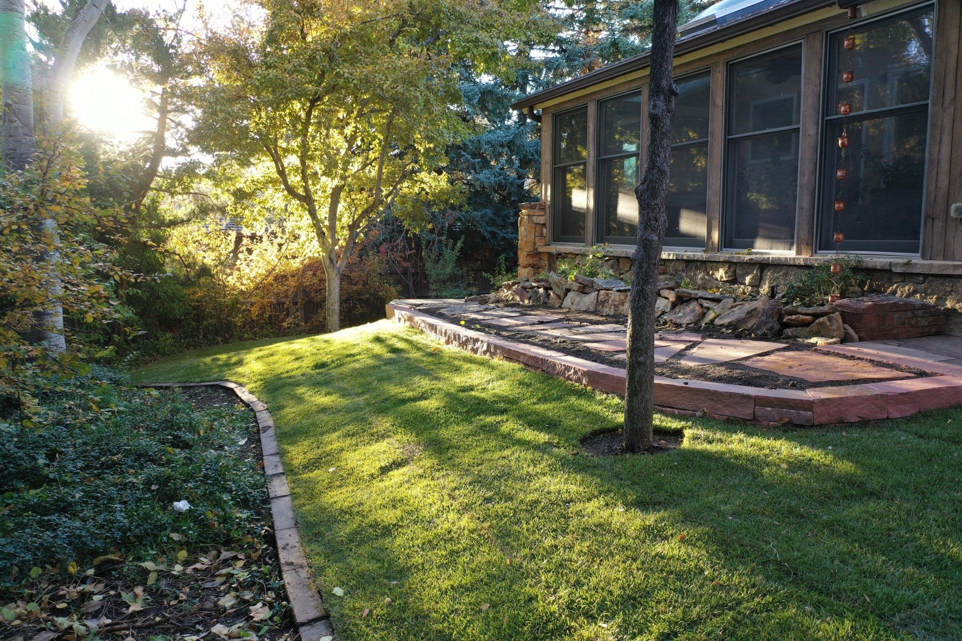 Sunlit backyard with grassy lawn, stone patio, and screened porch. Trees and shrubs surround.