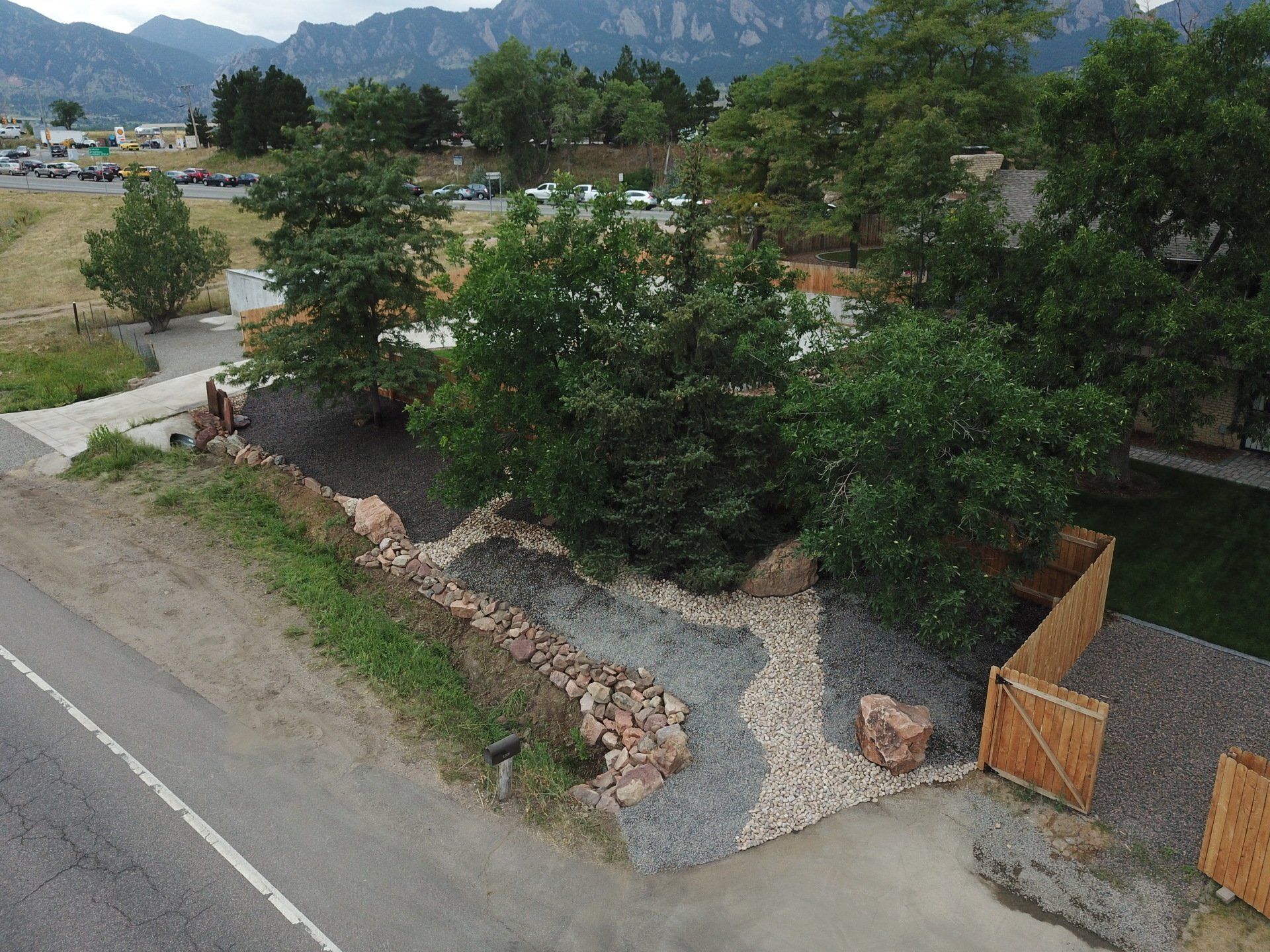 Landscaped yard with trees and gravel pathway next to a road, mountains in the background.