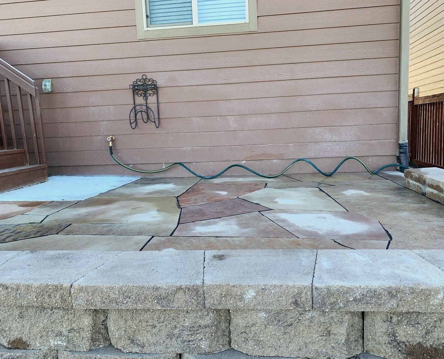 Patio with flagstone pavers, hose, and retaining wall against a light brown building.
