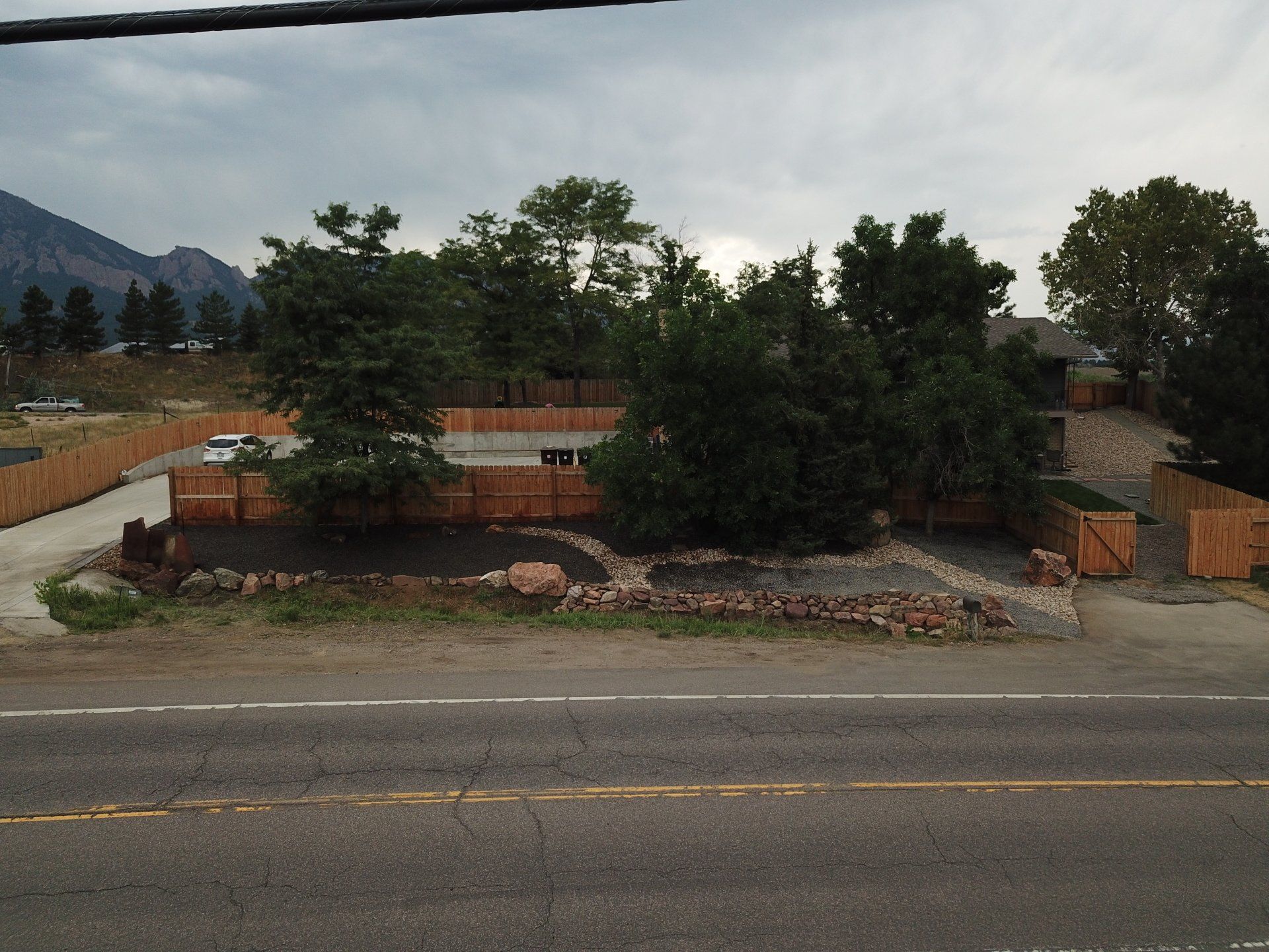 Brown wooden fence encloses parking area with trees and a dirt patch. Road in foreground, mountains in the distance.