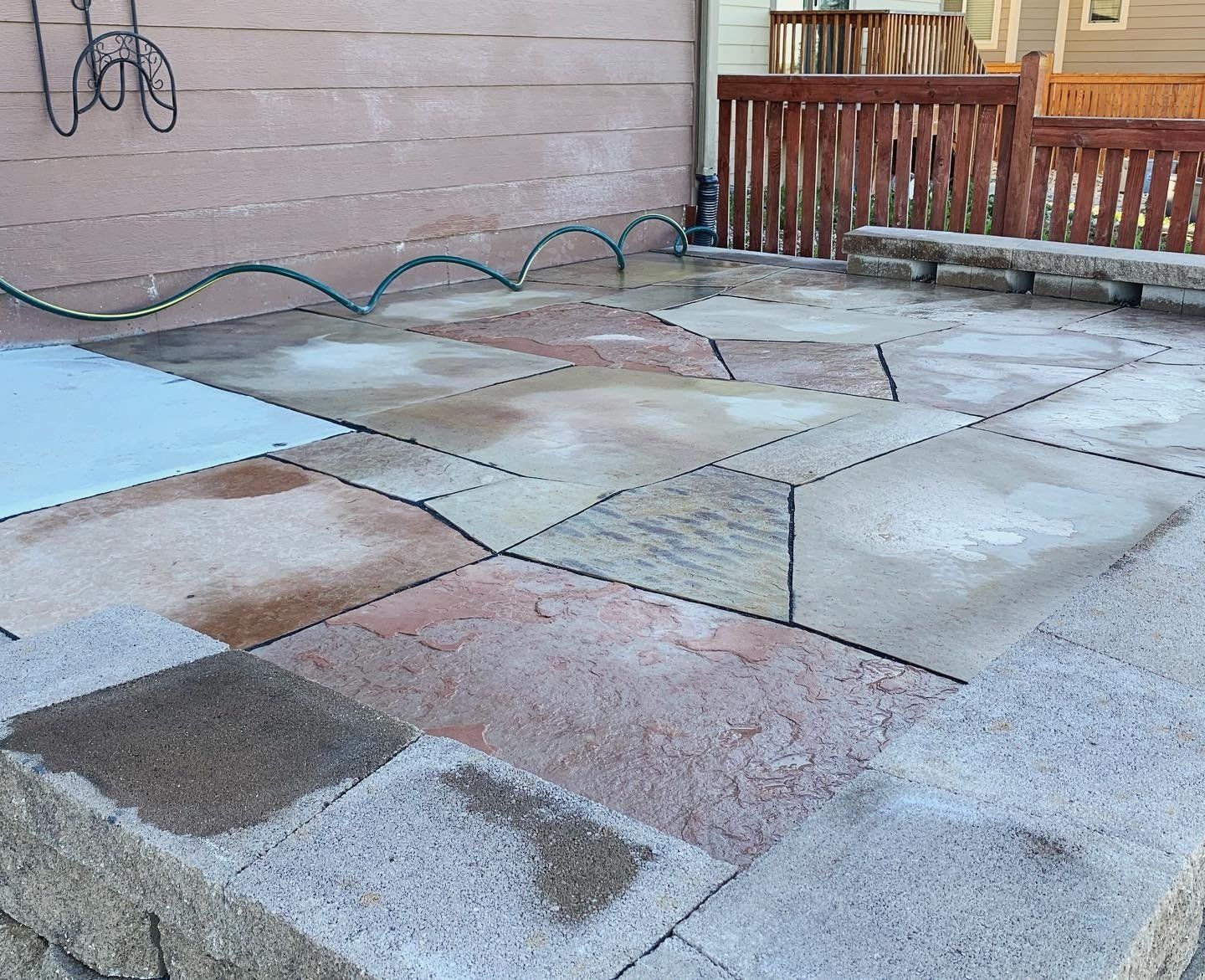 Patio with irregularly shaped stones, some wet, along a wall and a wooden deck. A hose arches over the patio.