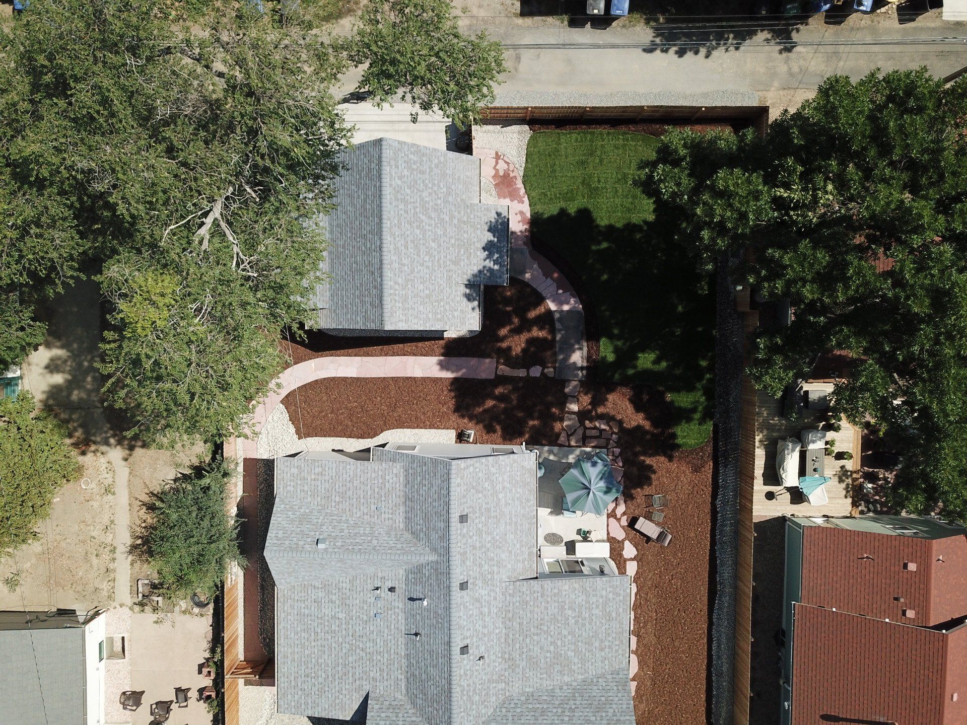 Aerial view of two houses with grey roofs, connected by a mulch path, surrounded by trees and green grass.