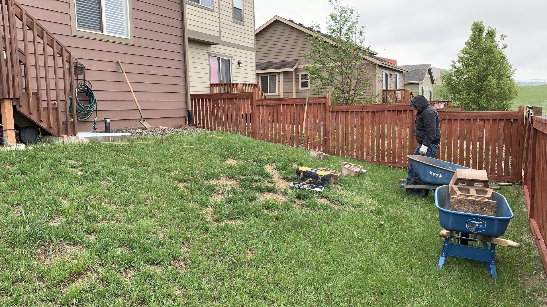 Person using wheelbarrows to move dirt in a grassy backyard, fenced with red wood.