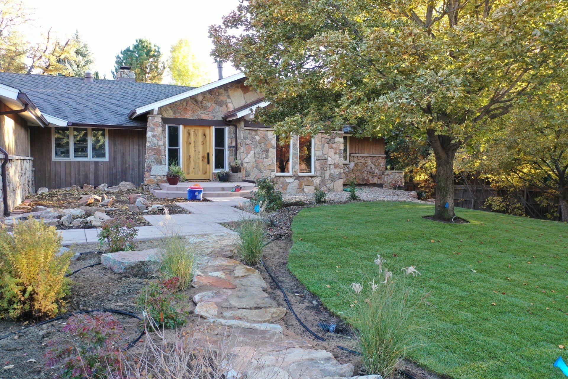 Stone-fronted house with stone walkway and green lawn, tree on right, autumn foliage.
