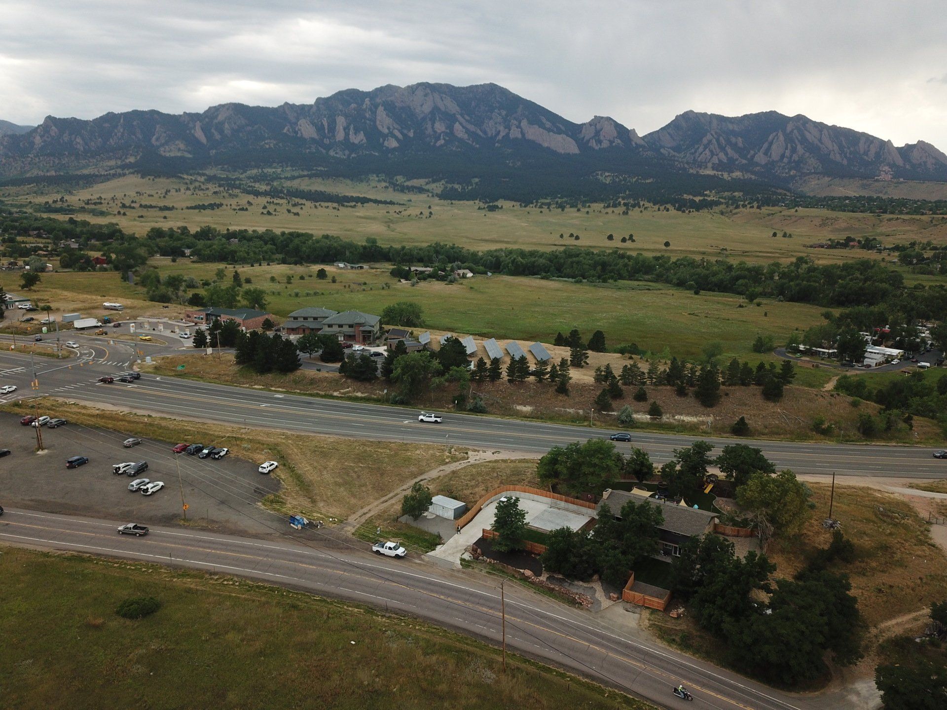 Mountains overlook a parking lot, buildings, and a road. Green fields and a cloudy sky surround.