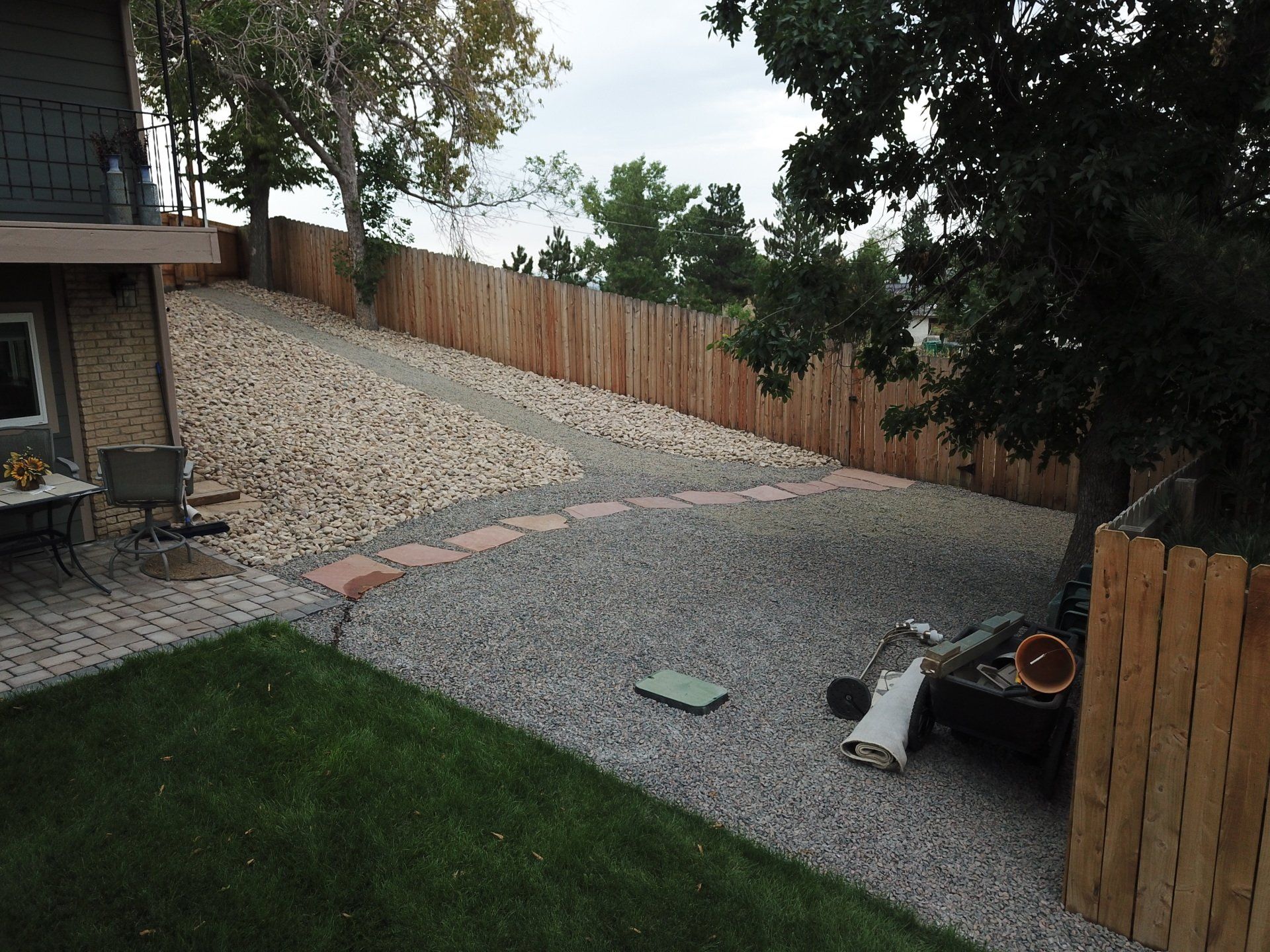 Backyard with sloped gravel area, wooden fence, grass, and a few trees.