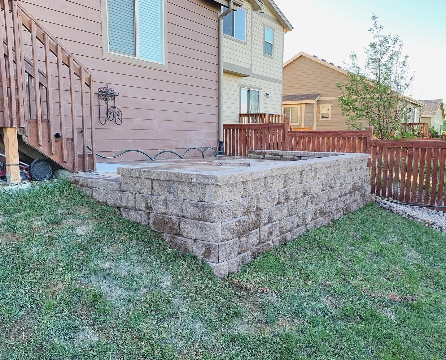 A stone retaining wall, bordering a patio and yard, next to a wooden deck and fence.